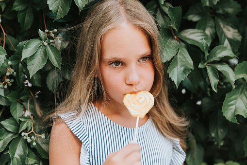 candid thoughtful little kid girl of eight years old holding lollipop and lick eating fruit sugar sweet candy on the background of a hedge wall of green plants in summer vacation outdoor
