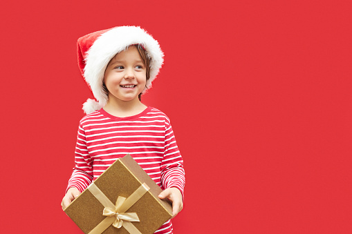 Charming boy in a santa claus hat and a gift box in his hands on a red background with space for text. New Year and Christmas gifts and sales.