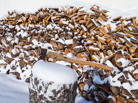 close up axe attached to the trunk of the tree on the background of piled chopped firewood covered with snow.