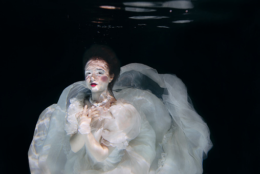 young woman in long white luxury dress underwater on the black background. Motion, art, winter concept