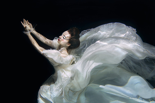 young woman in long white luxury dress underwater on the black background. Motion, art, winter concept