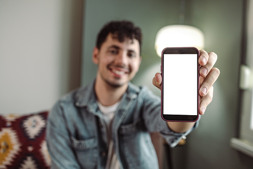 A young  man is at home, he is holding a white mobile phone screen and smiling