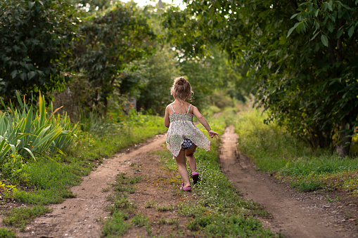 People. Children. A beautiful little caucasian light girl runs cheerfully and smiles along a country road in the summer in the village at sunset. Concept. Soft focus.