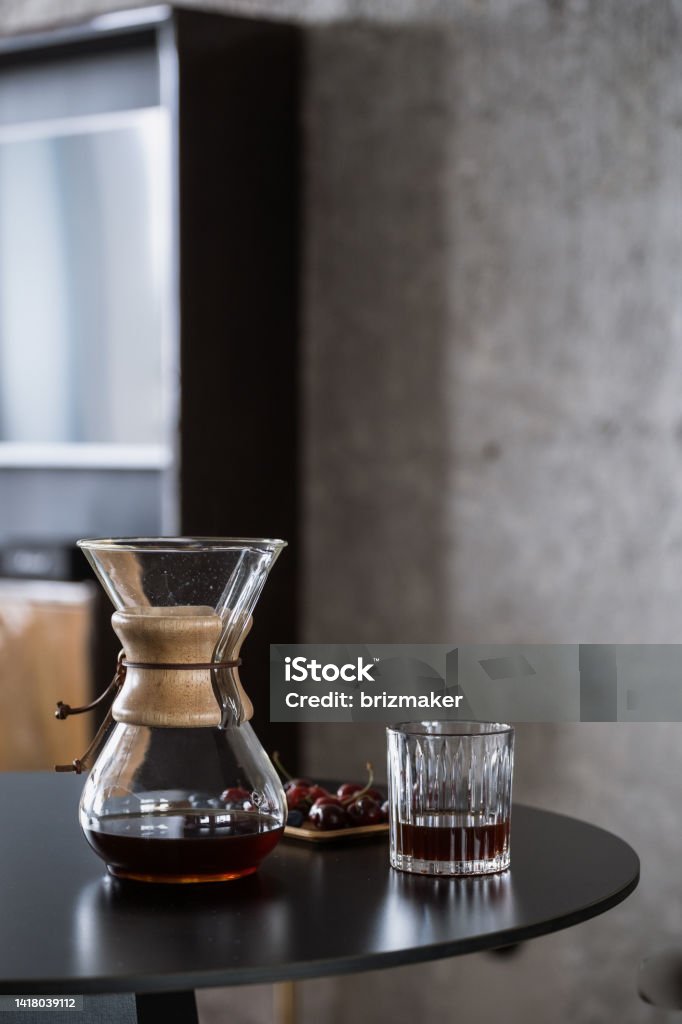 black filtered coffee in chemex and glass on table alternative method of brewing fresh coffee at home. glass chemex with hot brown caffeine beverage and wooden tray with sweet cherry on kitchen table Coffee - Drink Stock Photo