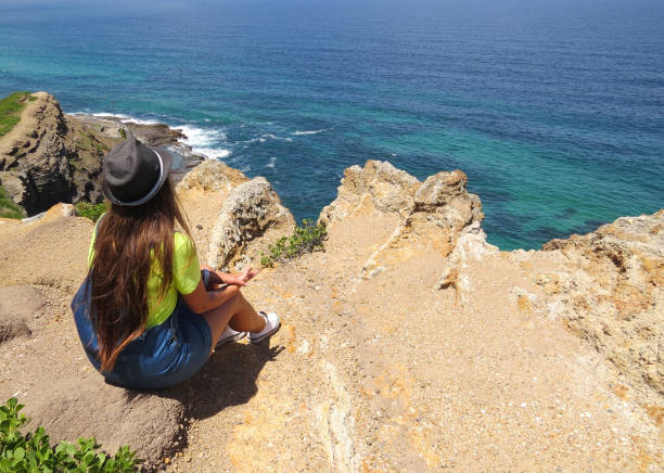 Girl with a hat and long brunette hair sitting on the cliff overlooking the blue ocean in Newcastle, Australia. stock photo
