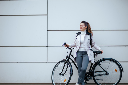 smiling stylish female in beige trench coat with bicycle In front of a metal wall outside on the city street.