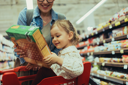 Happy young girl, sitting in a red shopping cart while her anonymous mother is pushing it. She is holding a box of cereal with excitement.
