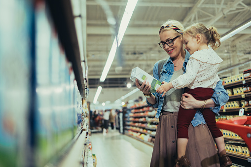Beautiful blonde women with glasses, wearing a denim jacket and a skirt, shopping with her young daughter in the supermarket. She is carrying the girl in her arms and picking up a box of milk or vegetable milk. The cute girl is curious while looking at the box, and mom is smiling.