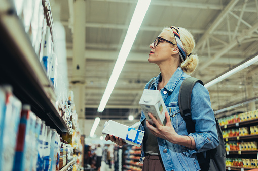 Beautiful blonde women wearing glasses, in a denim jacket and a skirt, shopping for milk in the supermarket. She is holding two cartons of milk and choosing which one to buy.