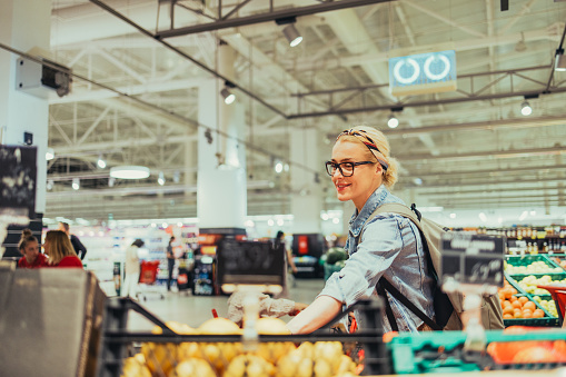 Beautiful cheerful blonde women, with her hair tied in a bun, wearing black glasses, denim jacket and a backpack, picking up fruit at the supermarket. She is looking down and smiling.