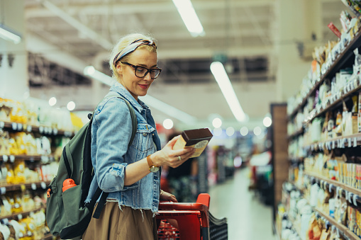 Beautiful blonde woman, pushing a shopping cart through the aisle and looking at products to buy. She is wearing glasses, a blue denim jacket and a backpack. She is holding and looking down at a product box, smiling while reading the ingredients.