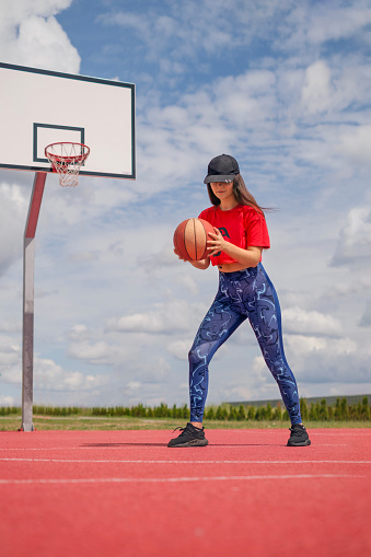A young girl is playing basketball on an outdoor playground with a red rubber coating on a hot sunny day. A person trains in a ball game