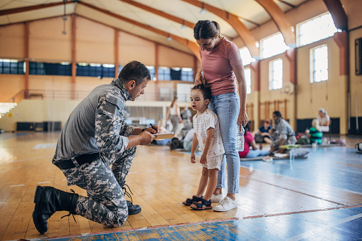 Diverse group of people, soldiers on humanitarian aid to civilians in school gymnasium, after natural disaster happened in city. Soldier talking with mother and son.mother