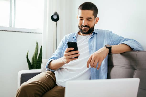 istock Smiling man sitting on couch at home using laptop and cell phone 1418058854