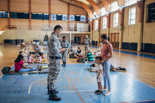 Diverse group of people, soldiers on humanitarian aid to civilians in school gymnasium, after natural disaster happened in city. Soldier talking with mother and son.