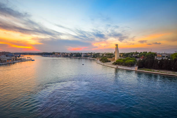 blick von einem kreuzfahrtschiff auf see auf das sailor monument und die hafenstadt brindisi, italien, in der südlichen region apulien unter einem bunten sonnenuntergangshimmel. - brindisi stock-fotos und bilder