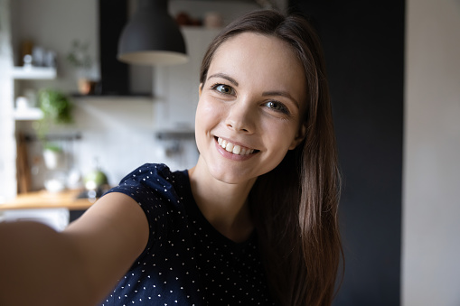 Selfie picture of happy beautiful young woman looking at camera with toothy smile. Pretty girl holding gadget with webcam, making video call, self home portrait. Communication concept