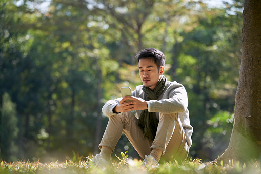 young asian adult man sitting on grass in park looking at cellphone