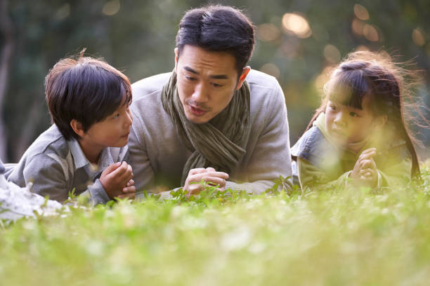 asian father and two children relaxing outdoors in city park stock photo