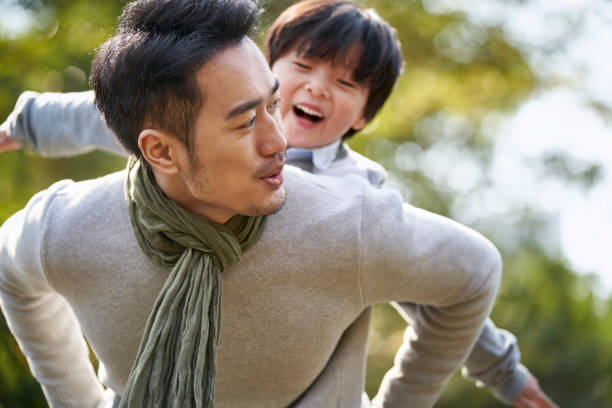 young asian father having fun carrying son on back outdoors in park stock photo