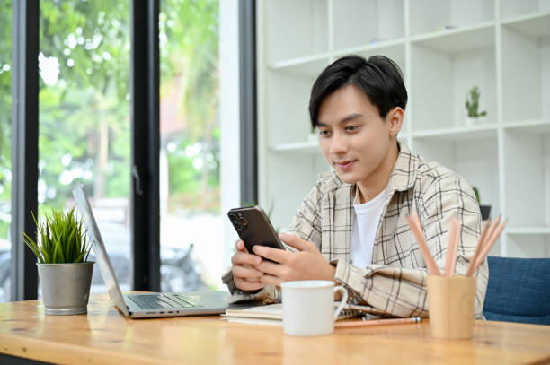 istock Cheerful Asian male at his office desk using smartphone to text, message, send email 1418085931
