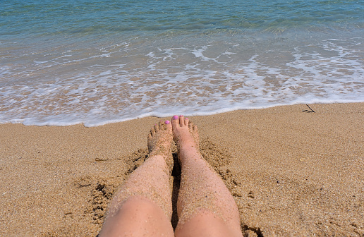 Women's feet with pink manicure in the sand on the beach against the backdrop of the waves and the sea
