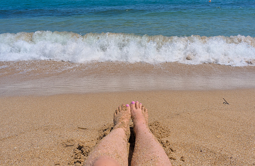Women's feet with pink manicure in the sand on the beach against the backdrop of the waves and the sea