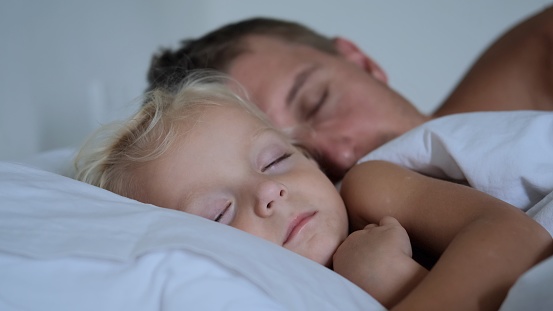 A young father, along with his daughter of two years, sleep soundly next to each other in bed on white bedding. They have deep dreams. They sleep in a bright cozy room. Father guards baby's sleep.