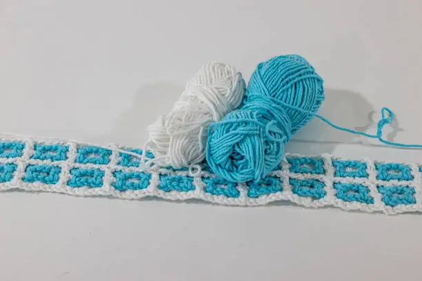 Interlaced crochet fabric in white and blue colors next to two balls of cotton against white surface and background, pattern in square shapes and in light relief. Handmade creativity