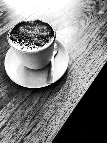 Vertical high angle closeup black and white photo of a ‘Liquid Gold’ drink: dark chocolate, black tahini, medicinal mushrooms and spices blended together and served hot in a ceramic mug on a wooden table in a cafe.
