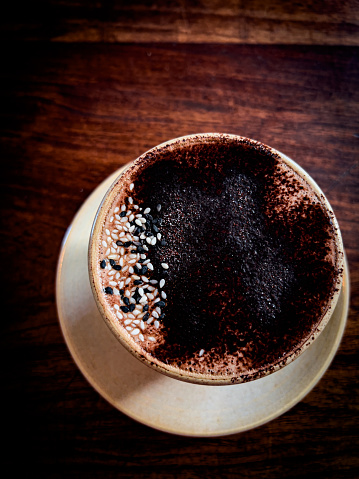 Vertical high angle closeup photo of a mug of ‘Liquid Gold’: dark chocolate, black tahini, medicinal mushrooms and spices, sprinkled with black and white sesame seeds and cocoa powder and served on a rustic wooden table in a cafe. Byron Bay, NSW.