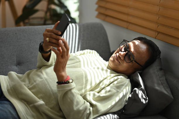 istock Happy middle age man chatting online in social network with smart phone while lying on couch at home. 1418122482