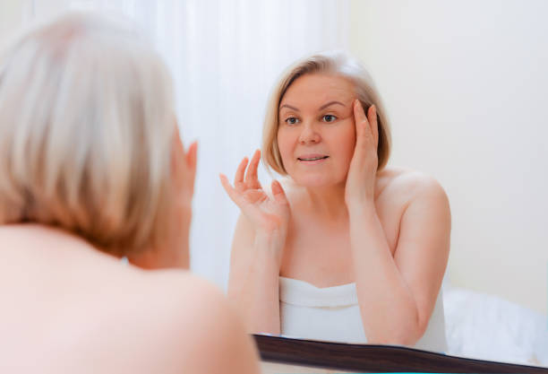 portrait pretty senior woman with hands on her face mirror at home after bathroom skin care after 50-60 years - make up women ceremonial makeup senior women imagens e fotografias de stock