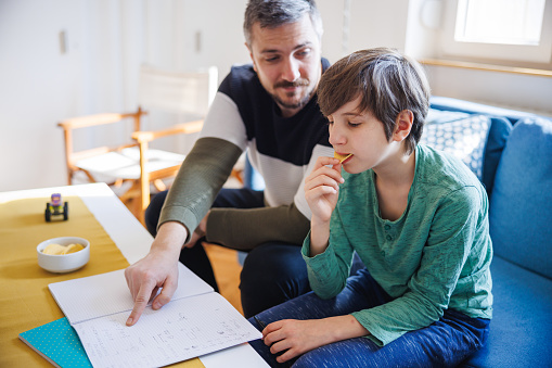 Father sitting next to his home schooled elementary aged son and pointing at task in notebook, the boy is having a snack