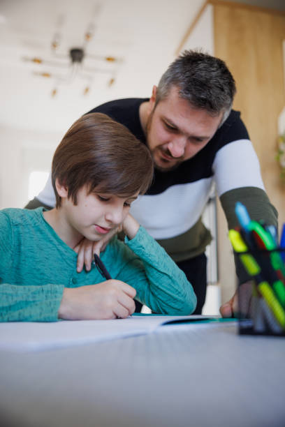 istock Tutor standing by home-schooled boy and looking at his notebook on the table 1418133764