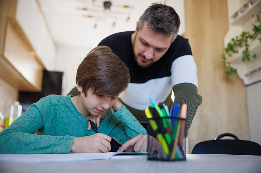 Elementary aged boy writing in his notebook, his tuition teacher standing by the table and pointing at the task