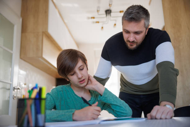 istock Tuition teacher standing by home-schooled boy and looking at his notebook on the table 1418133771