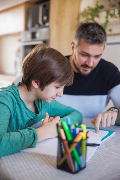 Tutor working with home-schooled boy and pointing at work in notebook stock photo