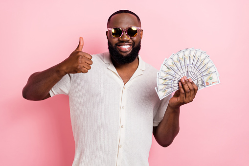 Photo of attractive confident guy dressed white shirt dark glasses holding money fan thumb up isolated pink color background.