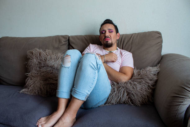 young man in make-up with a pained gesture of sad concern sitting on the sofa in his house. LGBTIQ people culture and rights young man in make-up with a pained gesture of sad concern sitting on the sofa in his house. LGBTIQ people culture and rights sad gay stock pictures, royalty-free photos & images