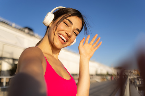 Portrait of a cheerful young adult sportswoman taking a break from her outdoor training session, having her wireless headphones on, looking at the camera and laughing
