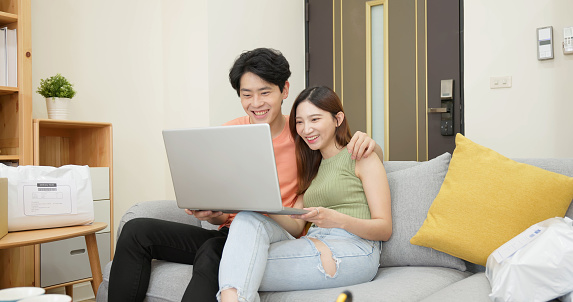 young asian couple sit on couch using laptop shopping together choosing goods buying online with many parcel in living room at home