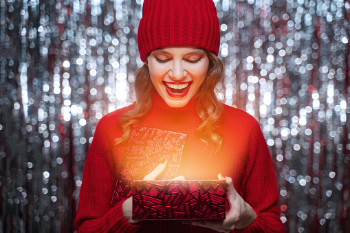 Christmas gift. Magic moment. Cute excite and surprised woman in warm red hat and sweater looking inside shiny present box on tinsel background. Happy new year. Merry xmas