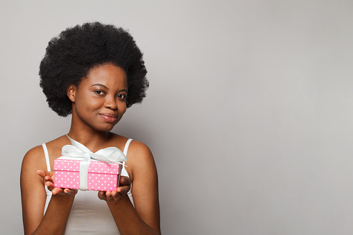 Cute cheerful black woman holding pink gift present box with white silky ribbon and looking at camera on white background