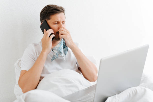 Sick man with flu symptoms using mobile phone and laptop while sitting on bed in bedroom. Sad ill guy holding handkerchief and calling on smartphone. Remote work on sick leave, cyberchondria concept stock photo