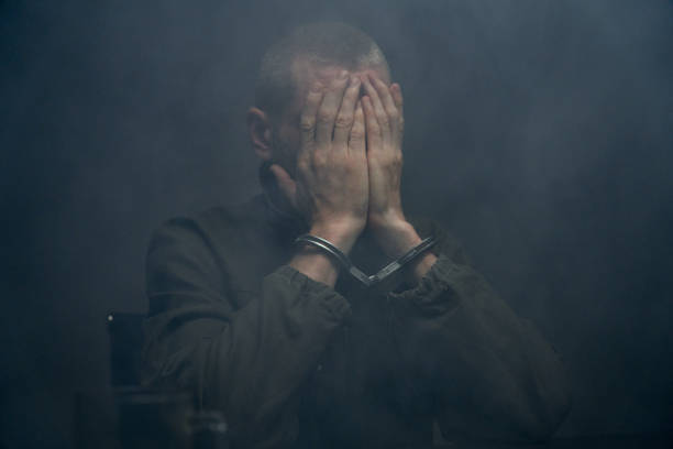 Close-up of a suspect in handcuffs closing his face with hands in a dark fogged room stock photo