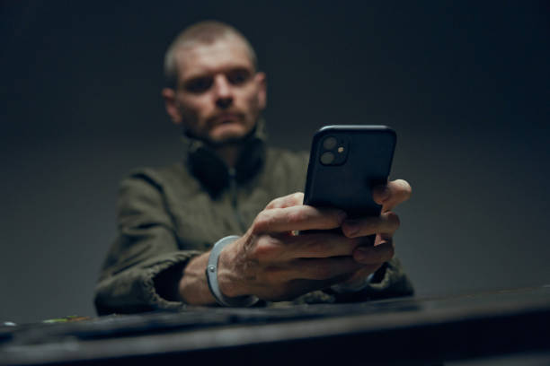 hands in handcuffs with a smartphone in the foreground, with a defocused background with a man, who's hands we see - prison cell imagens e fotografias de stock