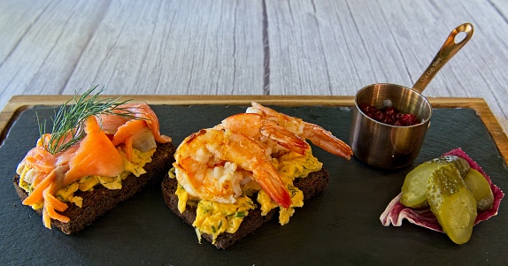 Two open sandwich, one topped with cold smoked salmon and scrambled eggs, another one topped with shrimps and scrambled eggs.