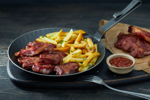 Grilled tasty ribs with French fries in an iron pan, with barbecue sauce on a dark wooden rustic background