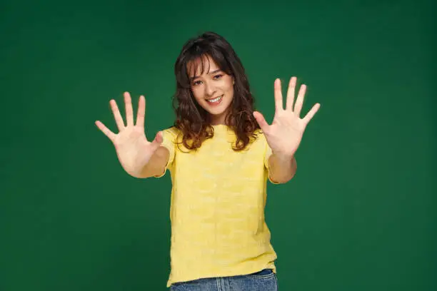 Friendly smiling young girl showing her palms, ten sign or stop gesture, outstretched atms, standing on green studio background. High quality photo